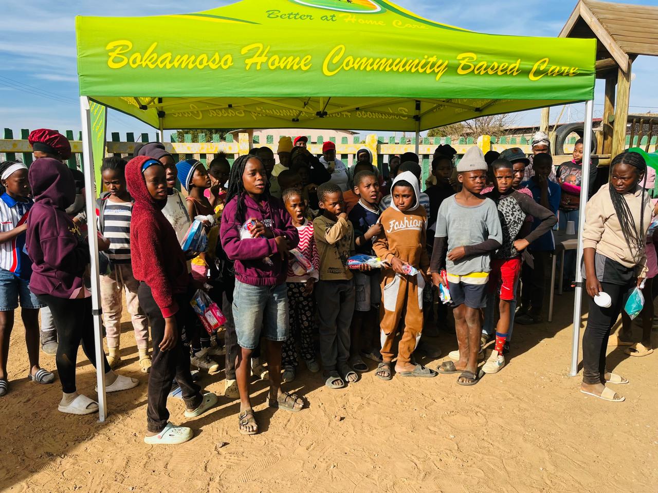 Children gathered under the Bokamoso canopy holding refreshments and supplies